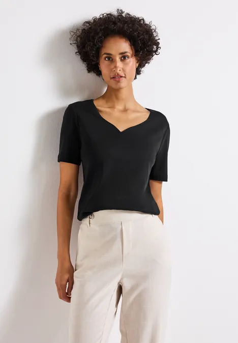 Frau mit schwarzem T-Shirt und heller Hose steht vor weißer Wand.